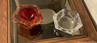 Photo showing two glass ashtrays: one clear faceted hexagonal and one amber with red center, also faceted hexagonal.