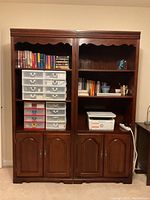 Front view of two large, dark wood bookcases with shelves and two-door cabinets at the bottom. Shelves hold plastic storage drawers, books, and other small items.