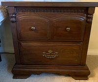 Close-up of one wooden bedside table showing two small upper drawers with round brass knobs and a larger bottom drawer with a decorative brass handle. Carved detailing on front and base visible.