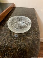 Round clear crystal ashtray placed on a textured brown granite surface, showing etched floral patterns around the rim with grooves for cigarette placement.