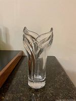 Clear crystal vase with a tulip-shaped top and decorative vertical leaf motifs on the sides, photographed on a dark surface against a plain wall.