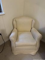 Front view of a small cream upholstered armchair placed against a wall, showing the full chair shape and cushion.