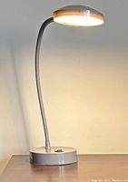 Photo of small desk lamp approximately one foot tall with flexible arm and round light head turned on, resting on a light-colored surface against light wall.