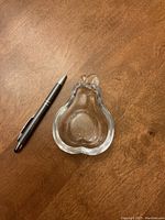 Clear glass pear-shaped dish next to a pen for size comparison on a wooden surface.