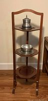 Photo of the small wooden display shelf with three tiers showing silver decorative items placed on the top and middle shelves. Shelf has a warm brown finish and curved top edge.