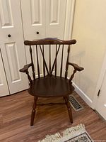 Front view of a dark varnished wooden Windsor-style chair with armrests, placed on a hardwood floor near white doors.