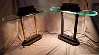 Two vintage halogen bankers desk lamps on soft fabric background, one black with brass columns, the other all black, both with UFO shaped glass shades