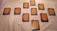 Photo shows 10 stacks of Magic the Gathering cards arranged on fabric surface. Each stack has roughly 50 cards. Card backs are visible with orange/brown design and MTG logo.