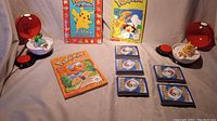 Front view showing all items: two Pokeball containers open with figurines (Pikachu and Bulbasaur), three Pokemon books arranged standing and laying flat, five stacks of Pokemon cards placed in the center