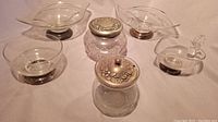 Two double sided candy dishes, small gravy/creamer pitcher, sugar bowl with silver lid, bowl with sterling base, crystal jar with lid displayed together on white surface.