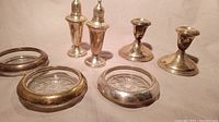 Photo showing two weighted sterling silver candlesticks, two sterling silver salt and pepper shakers, and three sterling silver ashtrays with crystal inserts arranged on a surface.