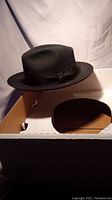 Black wool fedora hat displayed in its box with a curved brim and ribbon band