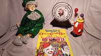 Four vintage circus clown items including a porcelain-faced clown puppet, a coloring book, a plastic clown toy, and a music box, arranged on a grey cloth background.
