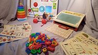Wide angle showing all items: Little Snoopy pull toy, ring stacking Color Stacks, School Days desk with chalkboard and letters, stencil sheets, and baby activity toys on a blue cloth background.