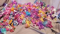 Wide assortment of approximately 100 vintage My Little Pony horses in varied colors and styles arranged on a beige fabric background.