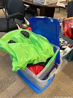 Adult green inflatable dinosaur costume partially unpacked from a blue plastic storage tote, showing the head and tail with black spots and some costume pieces inside the tote.