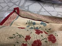 Close-up of folded area rug showing red and blue floral patterns, beige grid weave background and red border