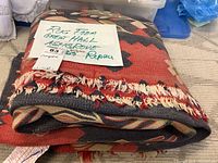 Folded rug showing geometric and floral pattern with damaged fringe edges and repair notes on attached tags.