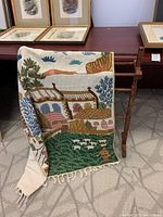 Handmade crewel tapestry hanging on the wooden blanket stand; tapestry shows colorful landscape with buildings, trees, and sheep.