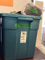 Blue plastic storage bin labeled with green tape indicating "x2 Curtain Panels" containing two folded floral patterned lined curtain panels and a clear plastic container with tassels labeled "Tassels".