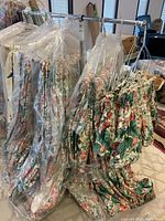 Side view of 6 floral printed curtains and valances hanging on clothes rack, some covered with plastic bags.