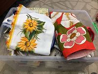 Top view of plastic container filled with dish towels including a white towel with hand-painted sunflowers and yellow crochet trim, and a red towel with large floral print.