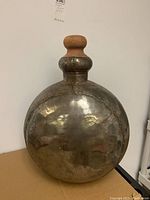 Front view of large steel flask with wooden knob top, showing surface dents and rust patches