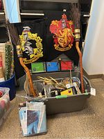 Photo showing two broomstick replicas positioned upright with natural bristle ends and wooden handles, in front of two large house flags for Gryffindor and Hufflepuff hung in the background. Several smaller boxed or packaged items and paper plates are arranged in a plastic tub below.