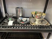 Shelf view of two clear glass dishes, cylindrical metal cooler, small plain bowl, ornate footed bowl on square tray