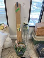 Full view of the boxed patio umbrella, metal tomato cage, and green ceramic planter with faux flowers and garden stakes in a room corner.