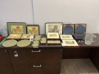 Overview of entire lot showing assorted Lady Clare placemats, clear glass pitcher, and two metal napkin rings on dark tabletop.