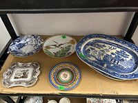 Six assorted plates and platters arranged on cardboard, showing various designs and color patterns including blue and white, multicolor, brown transferware; some with floral and Asian motifs.