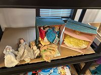 Seven porcelain dolls arranged on a shelf; three in original Madame Alexander doll boxes and four older dolls lying loose, showing vintage clothing and wear.