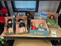 Seven Madame Alexander dolls in original boxes shown on table with window background. Dolls dressed in various colorful costumes.