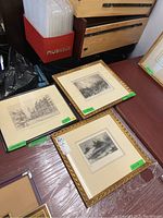 Three framed prints on a table, two in gold frames and one in a black frame with architectural and landscape scenes.
