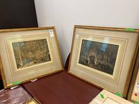 Two framed historic prints side by side on a dark wooden surface, showing architectural interiors with people and flags.