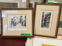 Two framed prints side by side on a table, showing detailed European street scenes in black and white, with handwritten notes and signatures.