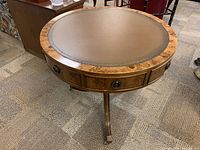 Angle view of the round wooden side table showing leather inset top, wood grain edge, and drawer with metal pull.