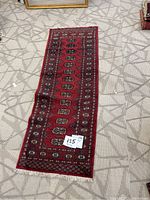 Full view of the red patterned runner rug laid out on a floor with beige geometric patterned carpet underneath, showing overall shape and design.