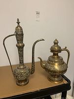 Photo showing both brass tea pots side by side on a flat surface, one tall and slender and the other shorter and rounder