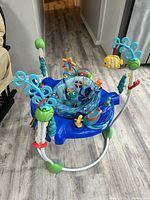 Photo of blue Baby Einstein kids jumper with attached colorful sea creature toys and padded fabric seat, placed on wooden floor.