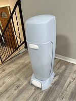Photo showing tall blue Diaper Genie diaper disposal system with a foot pedal, placed on wood floor next to stair rail and wall.