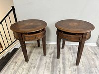 Two round wooden end tables with inlaid tops, drawers, and four legs, photographed against a white wall on a light grey wood floor.