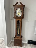 Full view of the tall wooden grandfather clock showing the entire case, glass door with visible pendulum and weights.