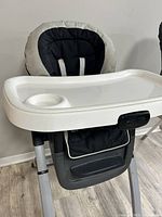 Graco high chair with white plastic tray including a cup holder and black/gray padded fabric seat with safety harness straps.