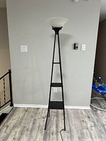 Full view of the black metal floor lamp with three triangular shelves and white bowl-shaped top shade against a wall.