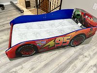 Side angle showing the Lightning McQueen toddler bed with mattress in place, graphics on the side panel include racing decals and bright red color with the number 95 and tire prints.
