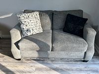 Photo of gray fabric loveseat with two accent pillows, white geometric pattern and solid black, placed in a room with light wood flooring.