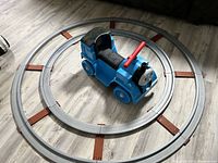 Full view showing the blue Thomas the Train ride-on toy centered inside the circular gray plastic track with brown accents on a wooden floor.