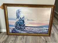 Framed print of steam locomotive train with scenic background in gold trimmed frame placed against a wall on the floor.
